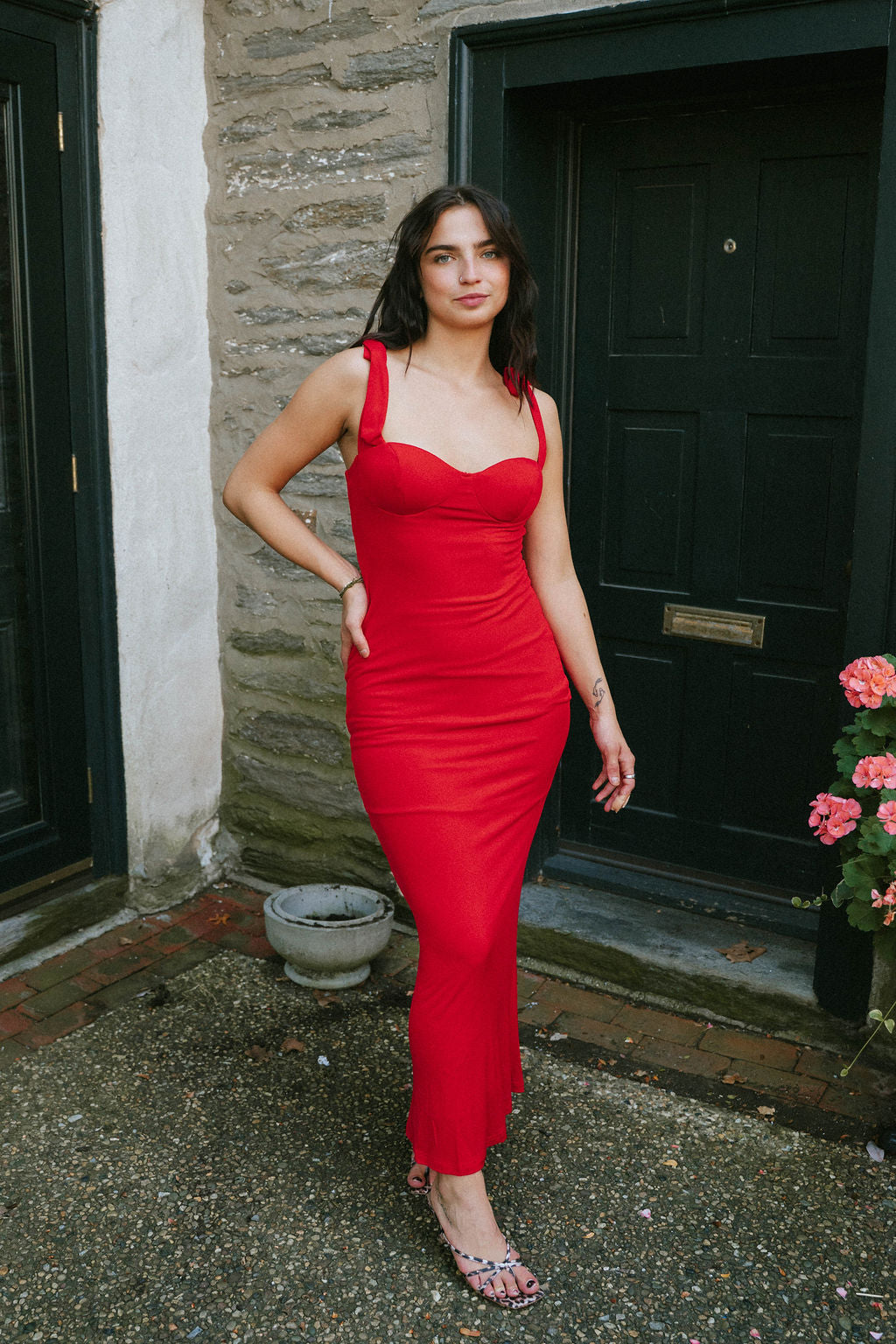 Woman wearing The Emilia Dress – Red, a fitted maxi with tie-shoulder straps and sweetheart neckline in bold red, styled with strappy heels.