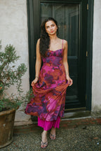 Woman wearing a vibrant floral maxi dress with thin straps in shades of magenta, plum, and rust, styled with strappy heels, standing by a dark door.
