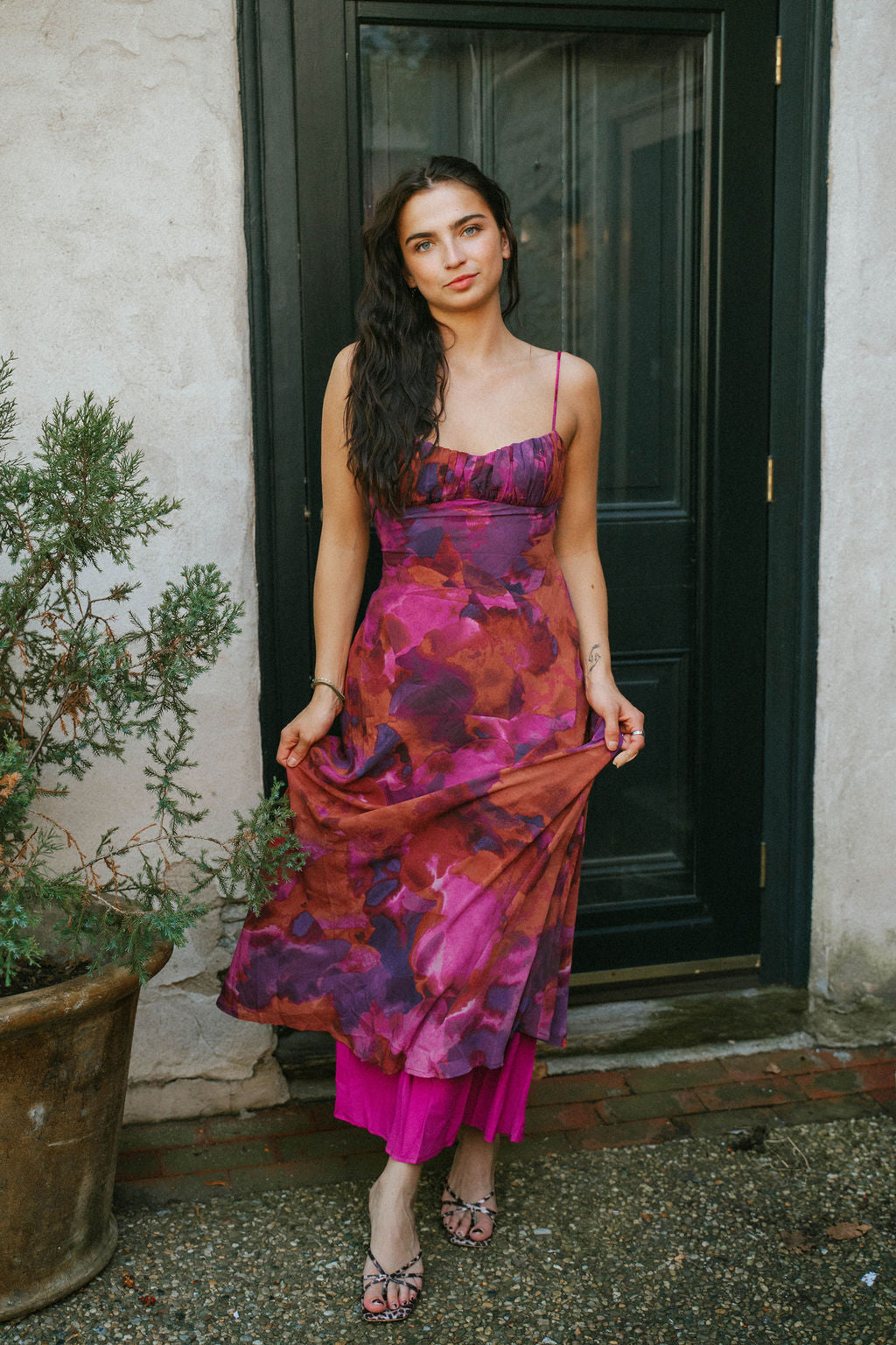 Woman wearing a vibrant floral maxi dress with thin straps in shades of magenta, plum, and rust, styled with strappy heels, standing by a dark door.